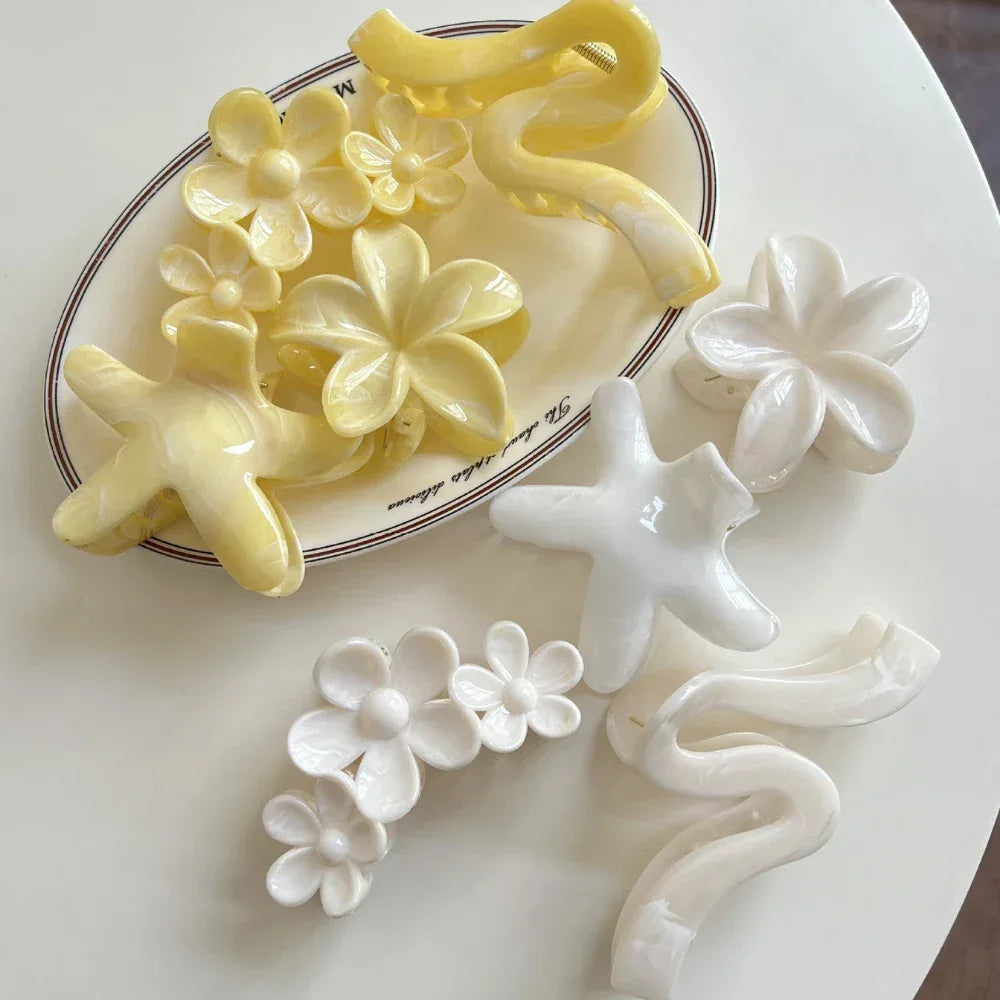 Various yellow and white hair accessories, such as flower clips, star clips, and wavy claw clips, are artfully arranged on a tray and a white surface