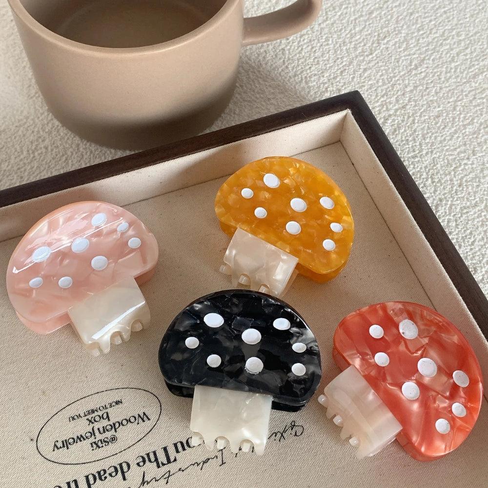 A collection of four mushroom-shaped hair claw clips in pink, orange, black, and yellow, arranged on a tray