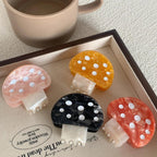 A collection of four mushroom-shaped hair claw clips in pink, orange, black, and yellow, arranged on a tray