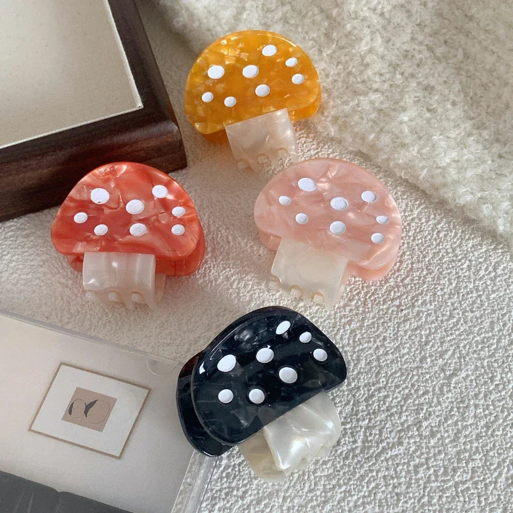Four mushroom-shaped hair claw clips in orange, pink, black, and yellow, arranged on a textured surface