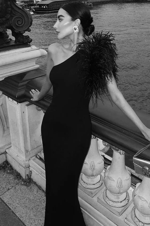 Black and white image of a woman in a black one-shoulder crepe gown with a feather accent on the shoulder and a mermaid hemline, posing on a balcony.