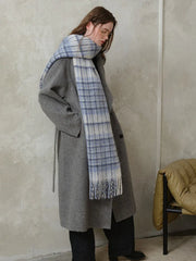 Woman standing with hands in pockets, wearing a grey coat and a blue and white plaid scarf.