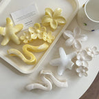 A collection of yellow and white hair accessories, including flower shapes, star shapes, and wavy claw clips, displayed on a white tray and table with a card and cup