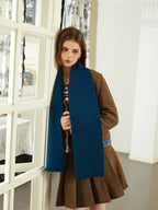 Model leaning against a doorway wearing a brown tweed jacket, denim shirt, blue scarf, and brown pleated skirt.