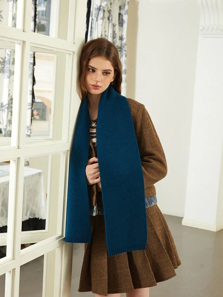 Model leaning against a doorway wearing a brown tweed jacket, denim shirt, blue scarf, and brown pleated skirt.