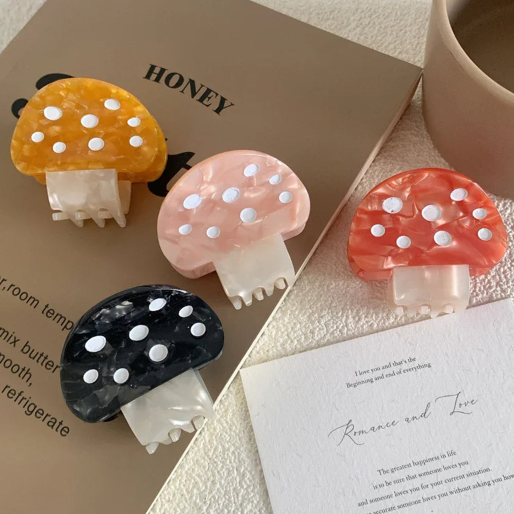 Four mushroom-shaped hair claw clips in orange, pink, black, and yellow, placed on top of books and stationery