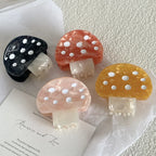 Four mushroom-shaped hair claw clips in black, orange, pink, and yellow, arranged on a white surface with stationery