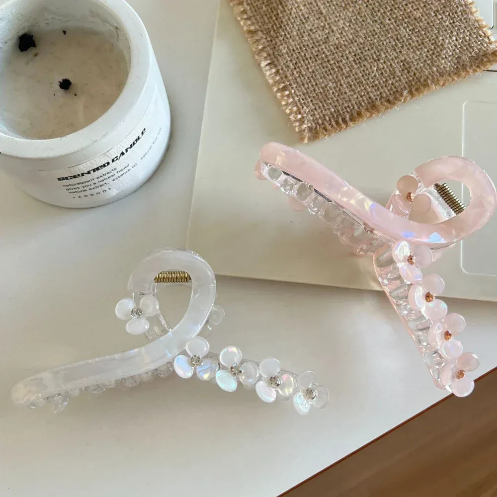 A white and a pink floral claw hair clip are showcased on a white table with a candle and a burlap square.