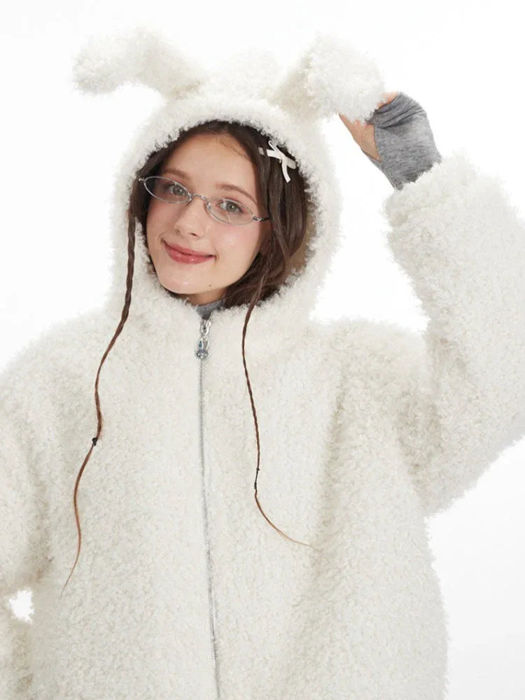 Close-up front view of a model wearing a white fuzzy hooded jacket with bunny ears pulled up. She is smiling and wearing glasses.