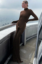 Woman wearing a long-sleeved, brown, mock neck, floor-length dress with a draped knot detail at the waist, posing on a balcony with a city view.