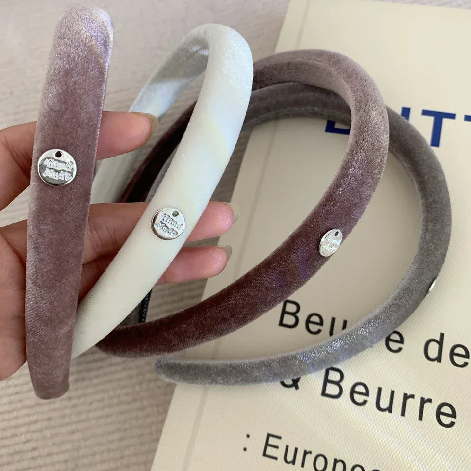 A hand holding three velvet padded headbands in taupe, cream, and grey, each with a silver charm. Secondary image