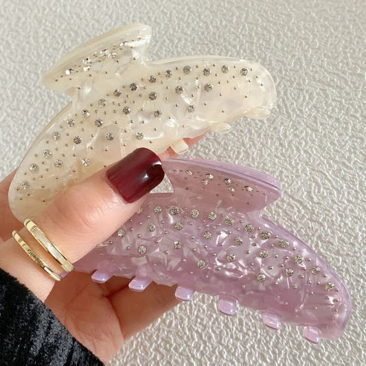 A hand holding two marbled claw clips, one cream and one lavender, both with rhinestones