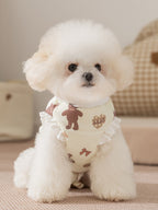 White poodle wearing a light yellow pet vest with a teddy bear print and ruffle trim, sitting down.