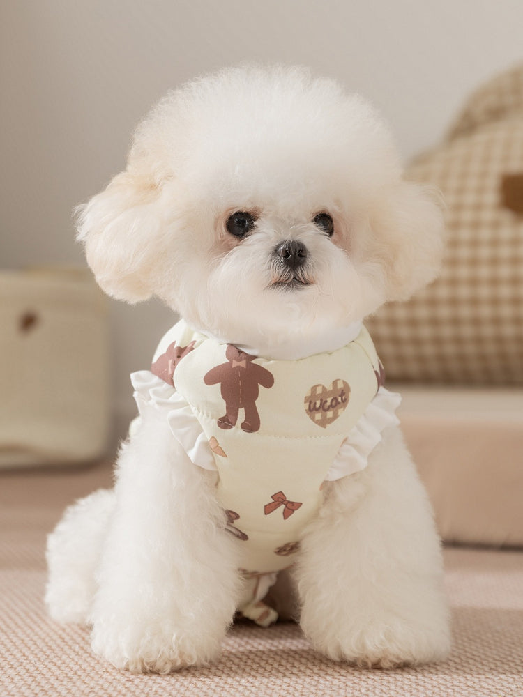 White poodle wearing a light yellow pet vest with a teddy bear print and ruffle trim, sitting down.
