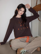 Young woman sitting on the floor wearing a brown long-sleeved graphic t-shirt and beige pants