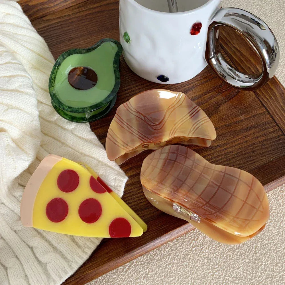 Several novelty hair clips, including a pizza slice, avocado, and peanut, arranged on a wooden tray.