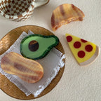 A top-down view of food-themed hair clips, including a pizza slice, avocado, and peanut, laid out on a textured surface.