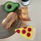 A flat lay of decorative hair clips, featuring a pizza slice, avocado, and peanut designs, alongside a mug and candle.