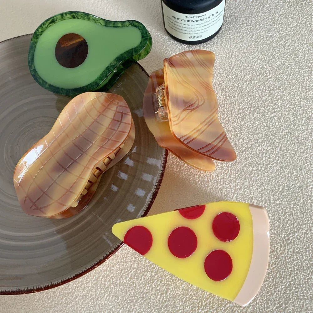 A flat lay of decorative hair clips, featuring a pizza slice, avocado, and peanut designs, alongside a mug and candle.
