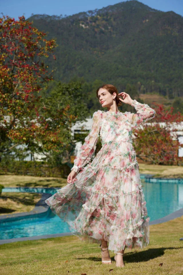 Woman posing in a flowing floral print chiffon gown with tiered ruffles and bishop sleeves, outdoors.
