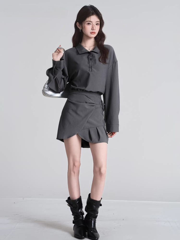 Model in a grey collared shirt and a pleated asymmetrical skirt, accessorized with black boots and a silver handbag.