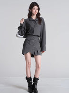 Model in a grey collared shirt and a pleated asymmetrical skirt, accessorized with black boots and a silver handbag.