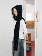 Woman wearing a black hooded scarf with a large bow detail, leaning against a wall.