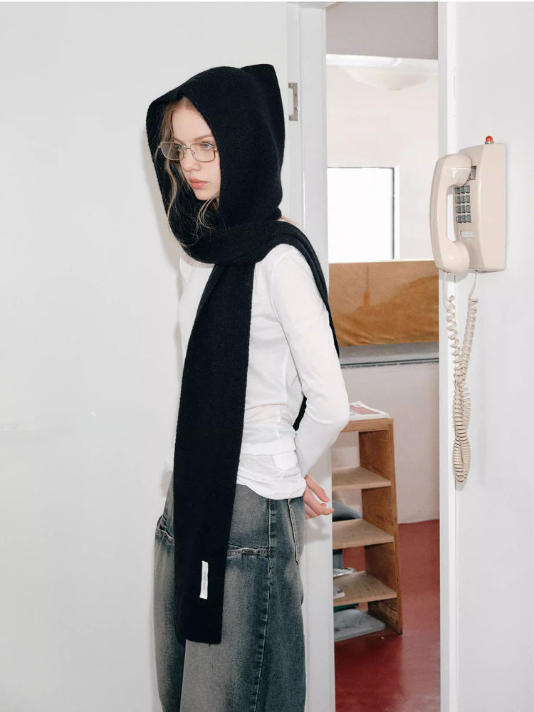 Woman wearing a black hooded scarf with a large bow detail, leaning against a wall.