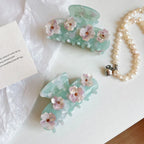 Two mint green acetate claw hair clips with floral embellishments, a small card, and a strand of pearls on a white surface.