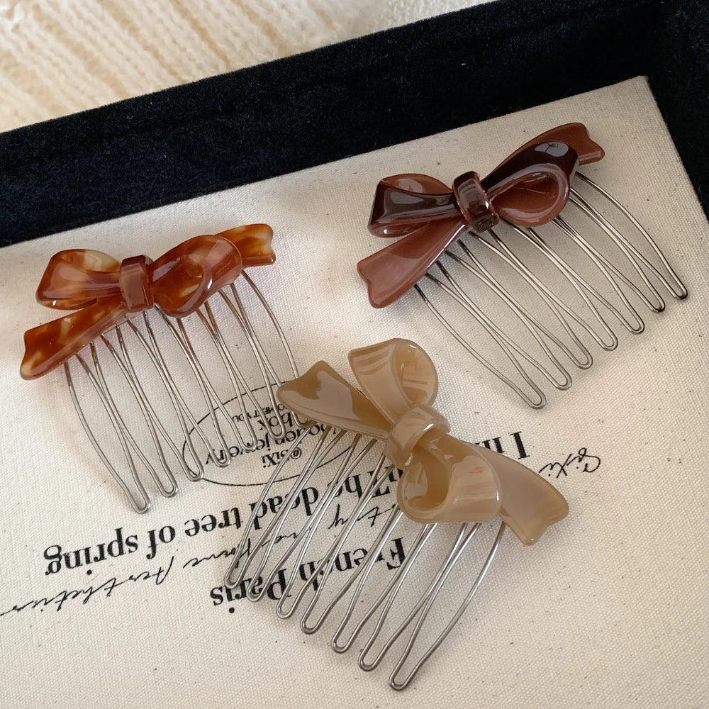 Three bow hair combs in brown, dark brown, and beige arranged on a textured white surface.