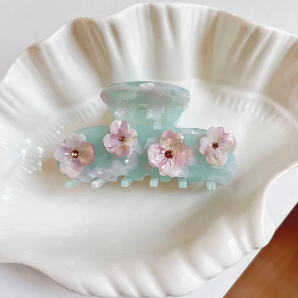 A mint green acetate claw hair clip with floral embellishments resting inside a white shell-shaped dish.