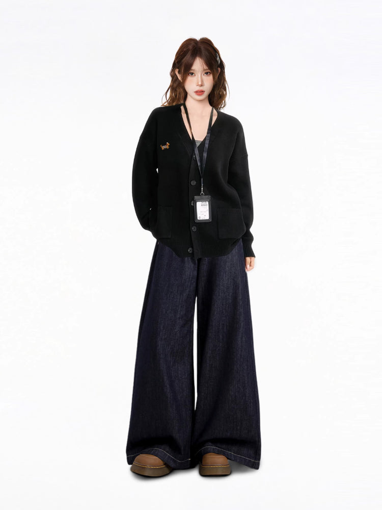 Full body shot of a model wearing a black V-neck knit cardigan with an embroidered dachshund detail, a black tank top, and dark wash wide-leg jeans, with an ID badge holder around her neck.