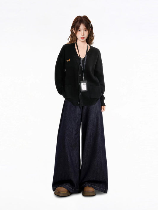 Full body shot of a model wearing a black V-neck knit cardigan with an embroidered dachshund detail, a black tank top, and dark wash wide-leg jeans, with an ID badge holder around her neck.