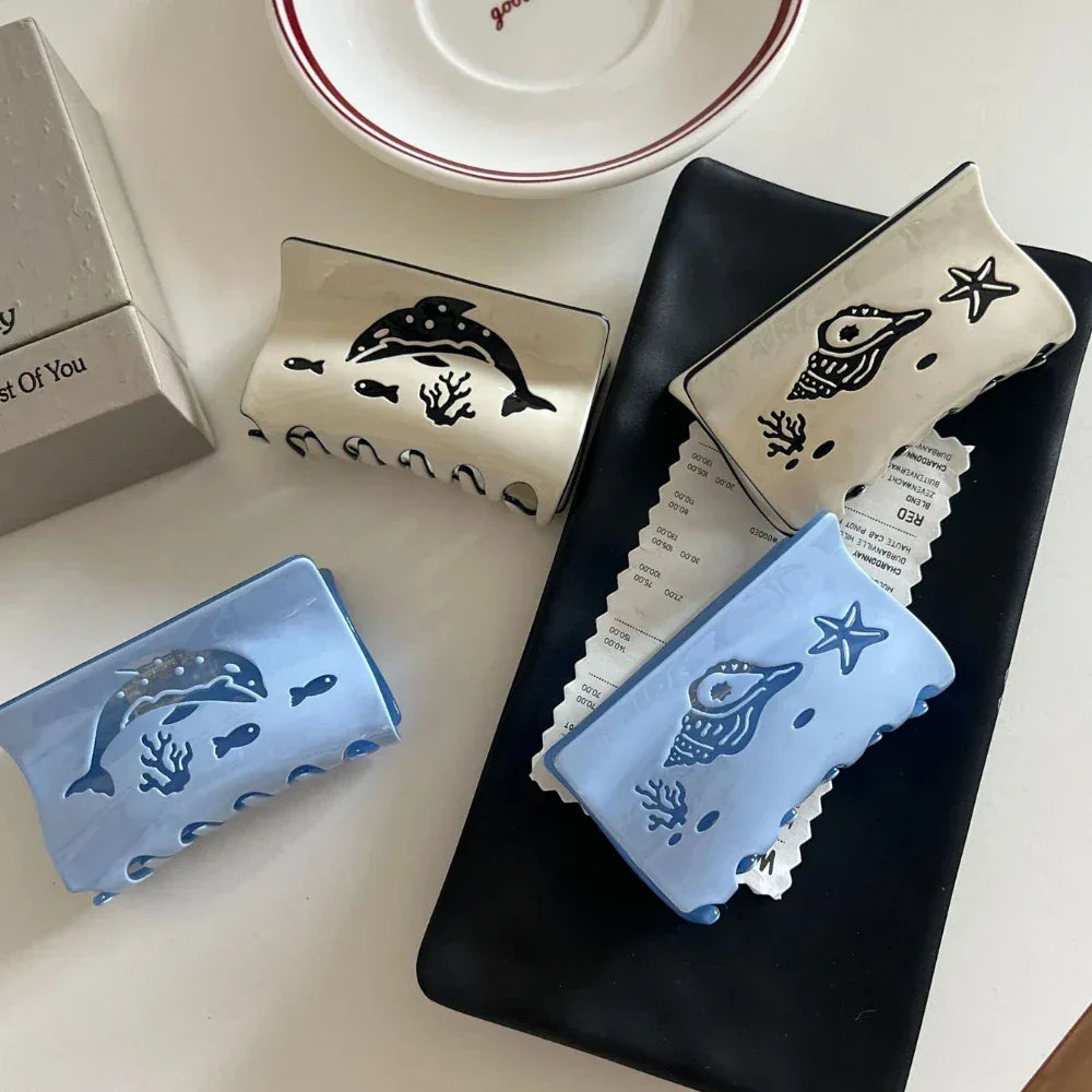 Collection of four hair claw clips with ocean-themed designs, including dolphins and seashells, in blue and cream colors