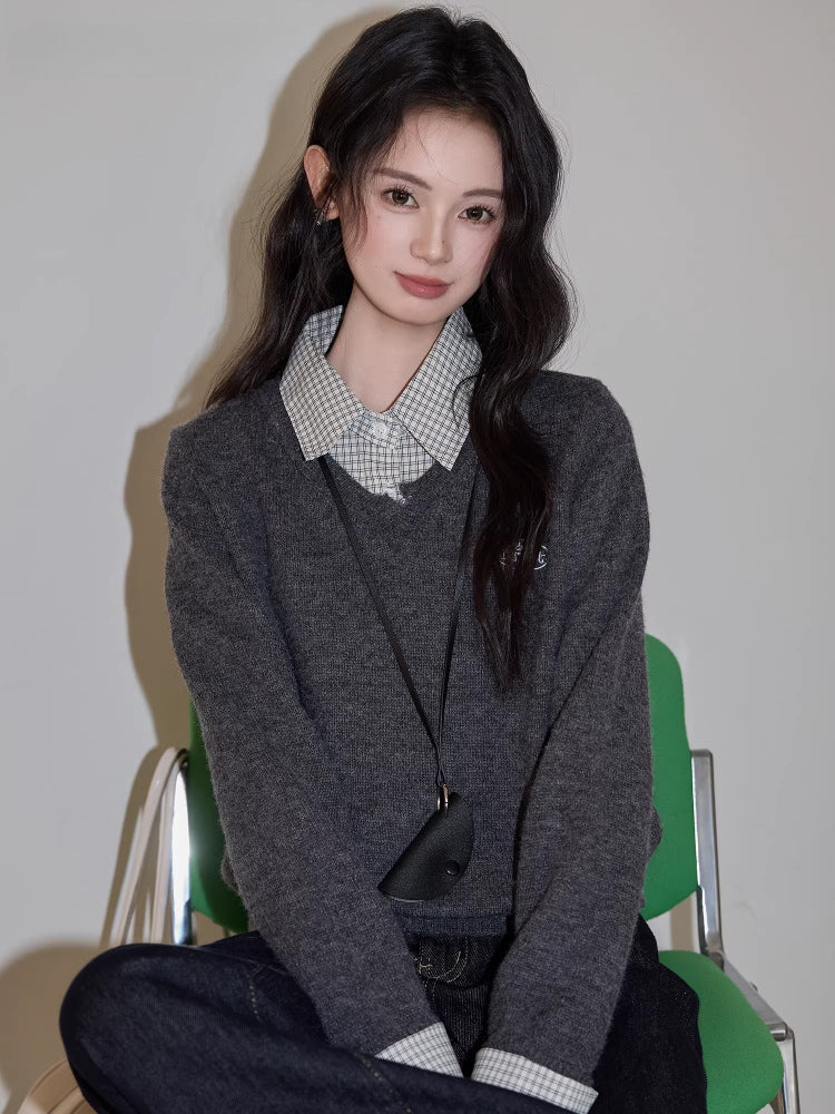 Close-up of a woman sitting on a green chair, wearing a grey v-neck sweater over a checkered collared shirt, dark wash wide leg jeans, and a black crossbody pouch.