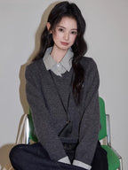 Close-up of a woman sitting on a green chair, wearing a grey v-neck sweater over a checkered collared shirt, dark wash wide leg jeans, and a black crossbody pouch.