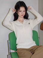 Woman sitting on a green chair, wearing a white cable knit henley sweater and brown pants, adjusting her hair.