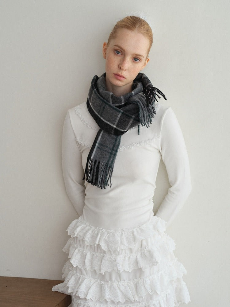 A young woman leans against a white wall, wearing a white long-sleeved top with lace trim, a white ruffled skirt, and a grey plaid scarf.