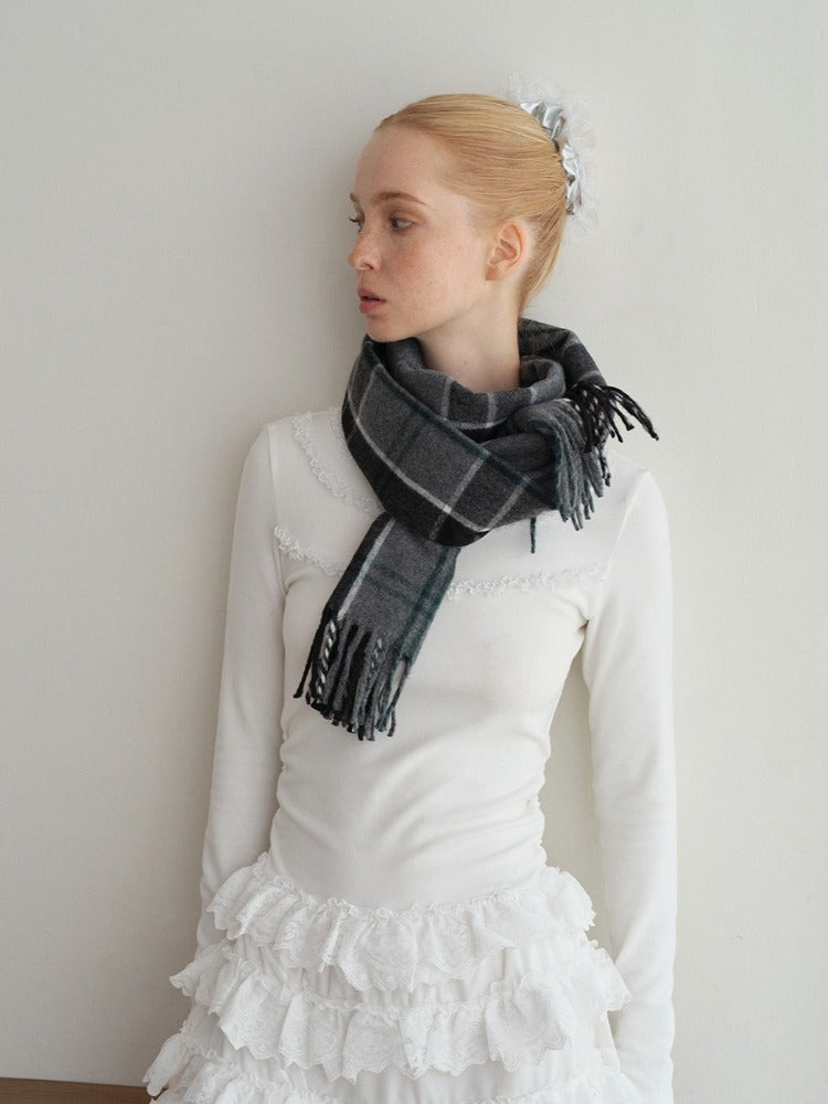 A young woman with her hair pulled back stands against a white wall, wearing a white long-sleeved top with lace trim, a white ruffled skirt, and a grey plaid scarf.
