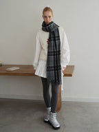 A young woman stands in front of a wooden table wearing a white long-sleeved shirt, a white vest with a floral design, a white pleated skirt, grey polka dot tights, silver boots, and a grey plaid scarf.