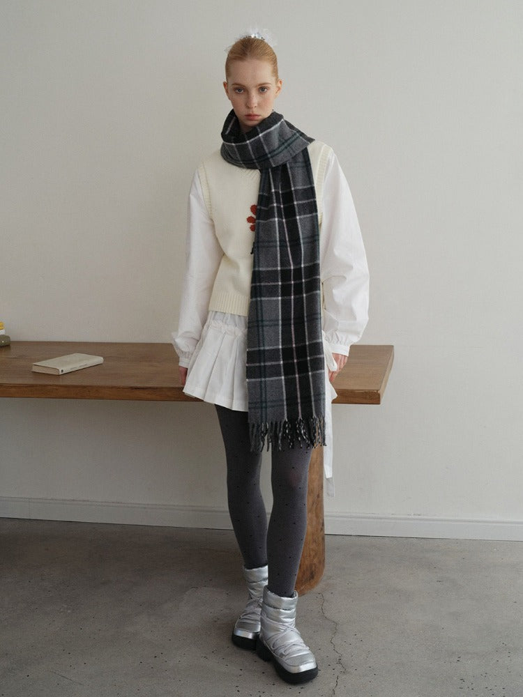 A young woman stands in front of a wooden table wearing a white long-sleeved shirt, a white vest with a floral design, a white pleated skirt, grey polka dot tights, silver boots, and a grey plaid scarf.
