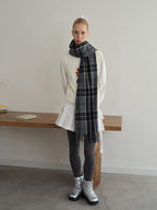 A young woman stands in front of a wooden table wearing a white long-sleeved shirt, a white vest with a floral design, a white pleated skirt, grey polka dot tights, silver boots, and a grey plaid scarf.