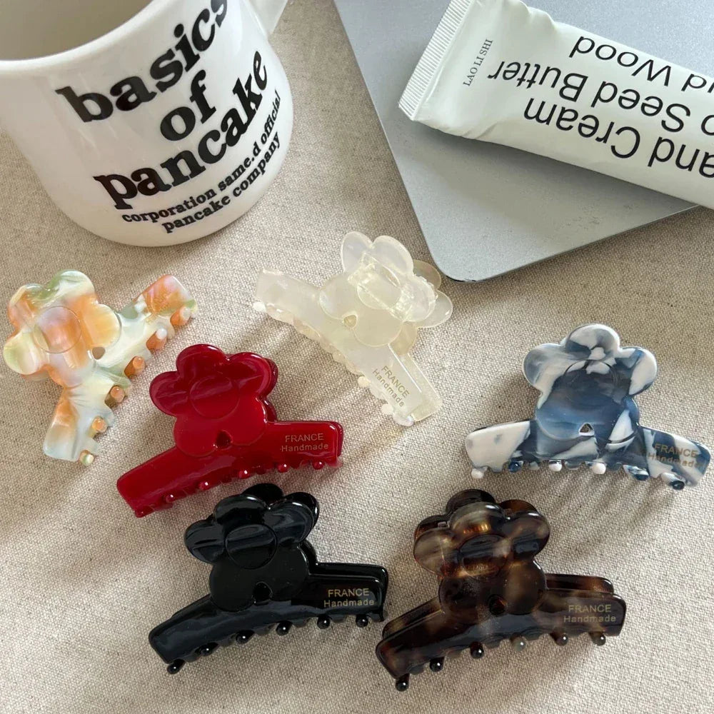 A collection of flower-shaped claw clips in various colors and patterns, including red, black, tortoiseshell, marbled blue and white, marbled orange and green, and translucent white, arranged on a textured surface next to a mug and a tube of hand cream.