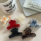 A collection of flower-shaped claw clips in various colors and patterns, including red, black, tortoiseshell, marbled blue and white, marbled orange and green, and translucent white, arranged on a textured surface next to a mug and a tube of hand cream.