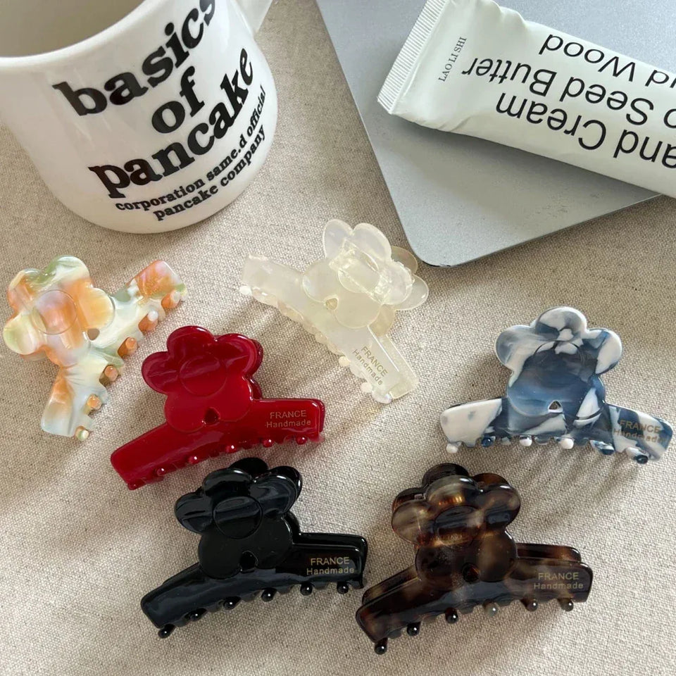 A collection of flower-shaped claw clips in various colors and patterns, including red, black, tortoiseshell, marbled blue and white, marbled orange and green, and translucent white, arranged on a textured surface next to a mug and a tube of hand cream. Secondary image