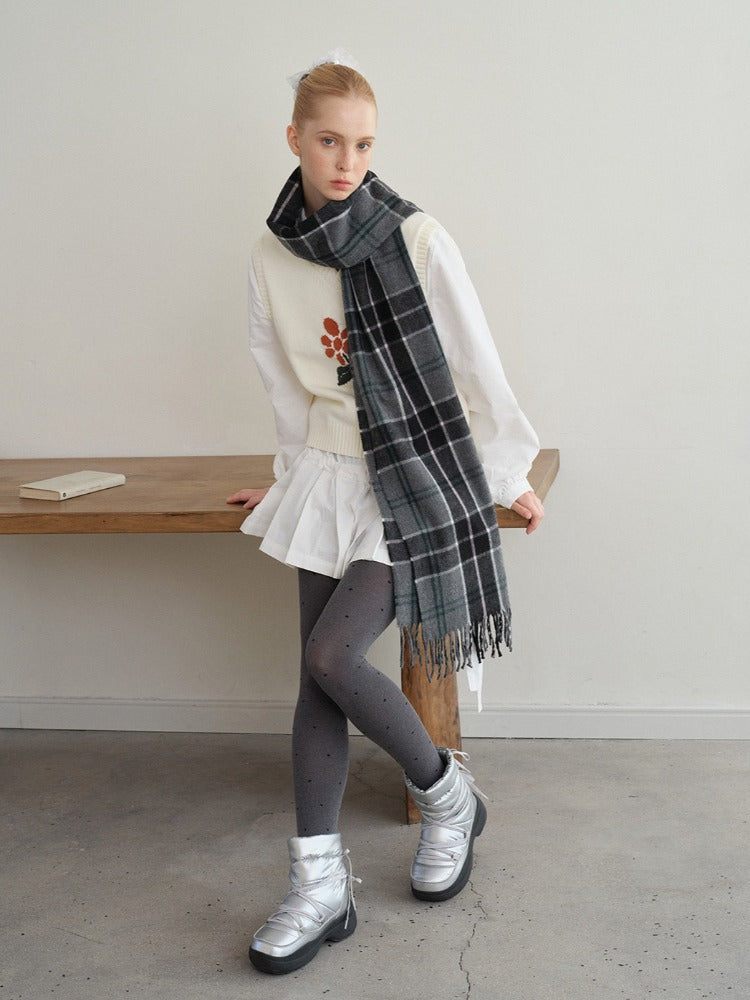 A young woman poses leaning on a wooden table while wearing a white long-sleeved shirt, a white vest with a floral design, a white pleated skirt, grey polka dot tights, silver boots, and a grey plaid scarf.
