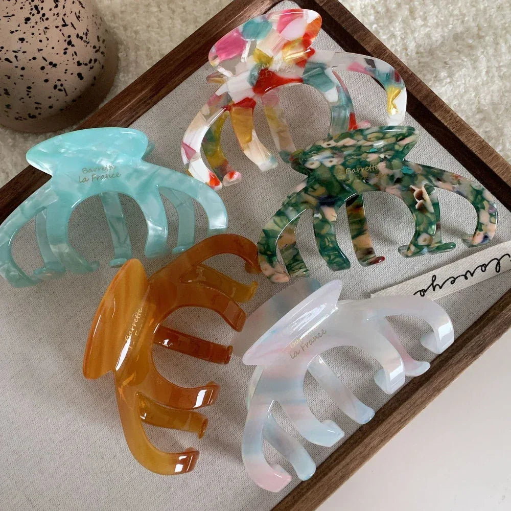 A collection of five acetate claw hair clips in various marbled colors: multi-colored, light blue, green, amber, and white with pink/blue accents. They are arranged on a wooden tray.