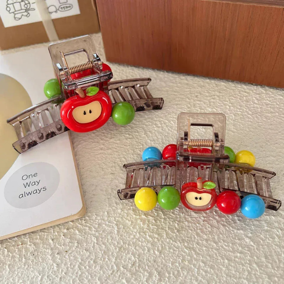 Two transparent hair claw clips, each with a red apple charm and colorful beads, placed on a textured surface next to a book. Secondary image