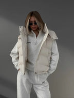 Woman wearing a cream puffer vest over a cream quarter-zip hoodie and cream joggers, with sunglasses and hands in pockets.