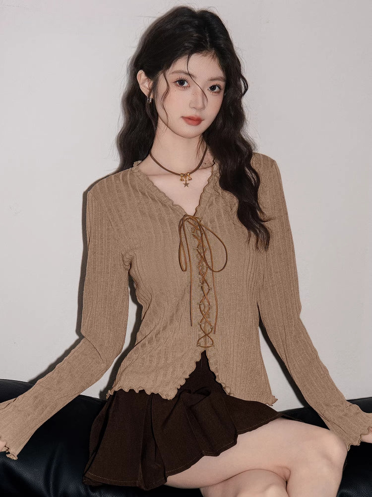 Woman sitting and wearing a tan ribbed knit long-sleeve top with a tie-front closure and ruffled hem, paired with a brown pleated skirt.